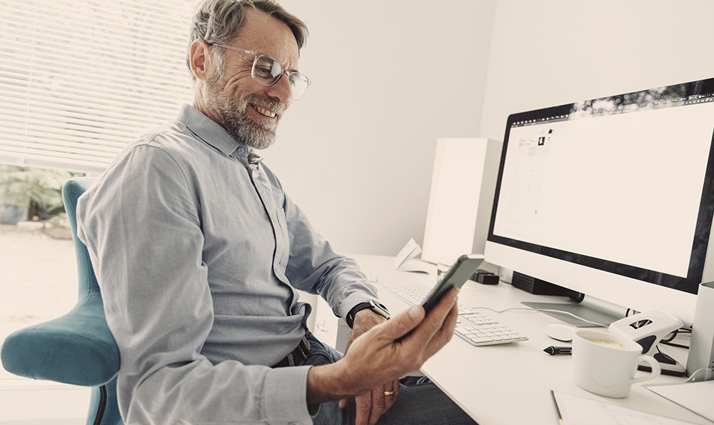Ein Mann mit grauen Haaren, Bart und Brille sitzt vor seinem Computer am Schreibtisch und schaut lächelnd in sein Smartphone