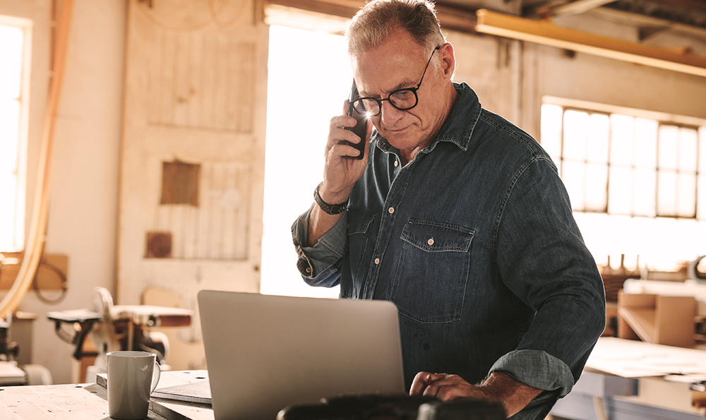 Ein Mann in einem Jeanshemd und mit Brille steht in einer Werkstatt, telefoniert und schaut dabei auf einen Laptop.