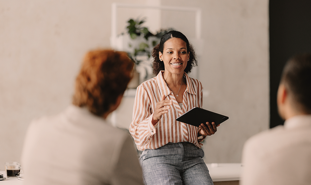 Eine Frau mit einem Tablet unterhält sich mit zwei sitzenden Personen. Sie lächelt, trägt ein gestreiftes Hemd und Jeans, im Hintergrund ist eine Pflanze zu sehen, was eine freundliche und professionelle Atmosphäre vermittelt.