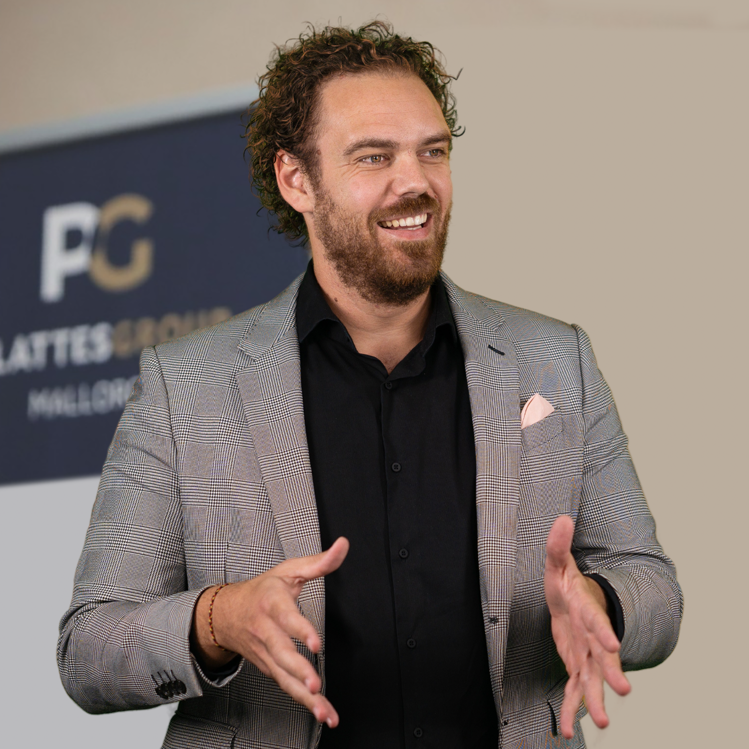 Christian Plattes mit lockigem Haar und Bart, in einem grauen Anzug und schwarzem Hemd, lächelnd und gestikulierend. Im Hintergrund ist ein unscharfes Schild mit dem „PG“-Logo zu sehen.