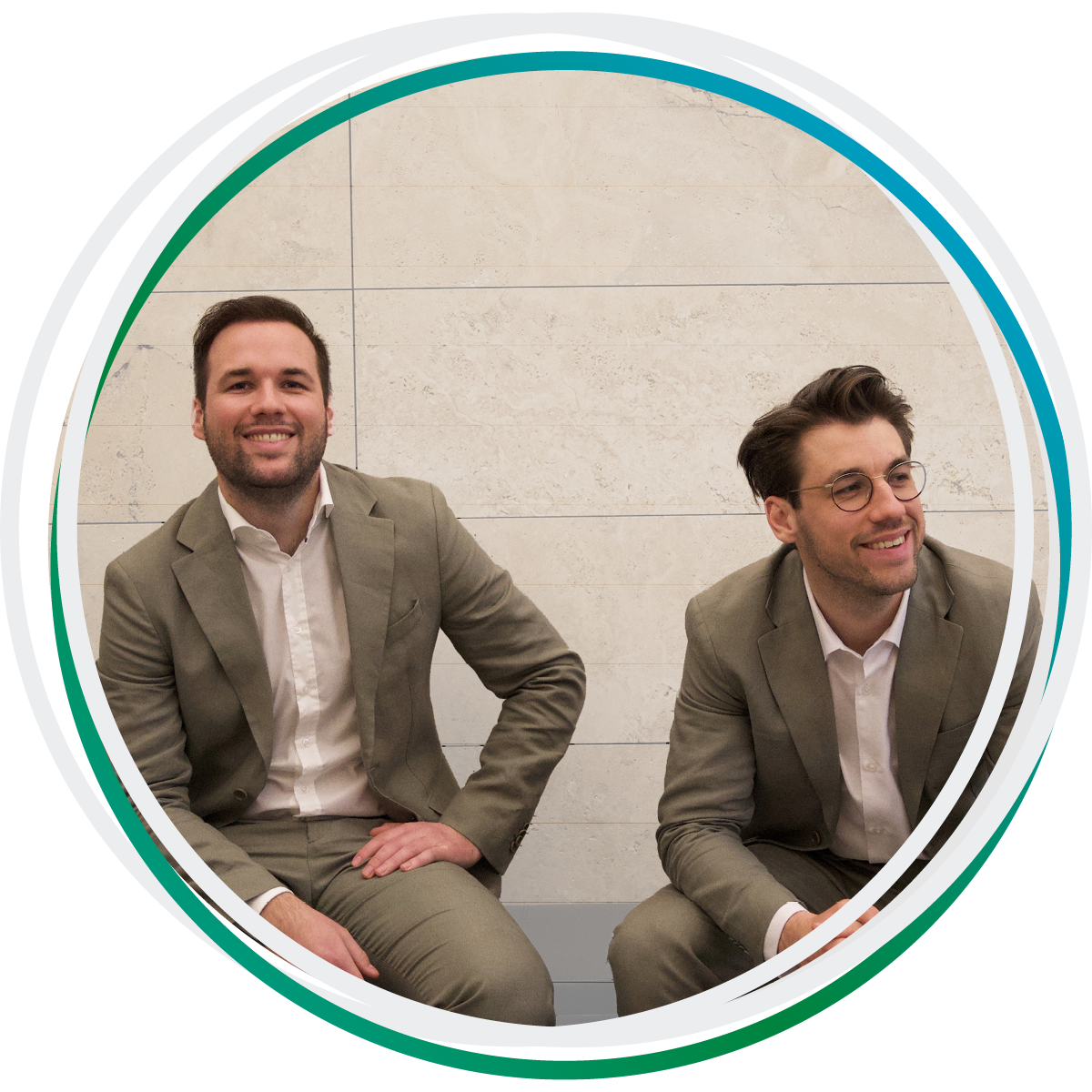 Die Rechtsanwälte Niklas und Lukas Hufeld sitzen vor einer Mauer und lächeln. Beide tragen einen beigen Anzug.