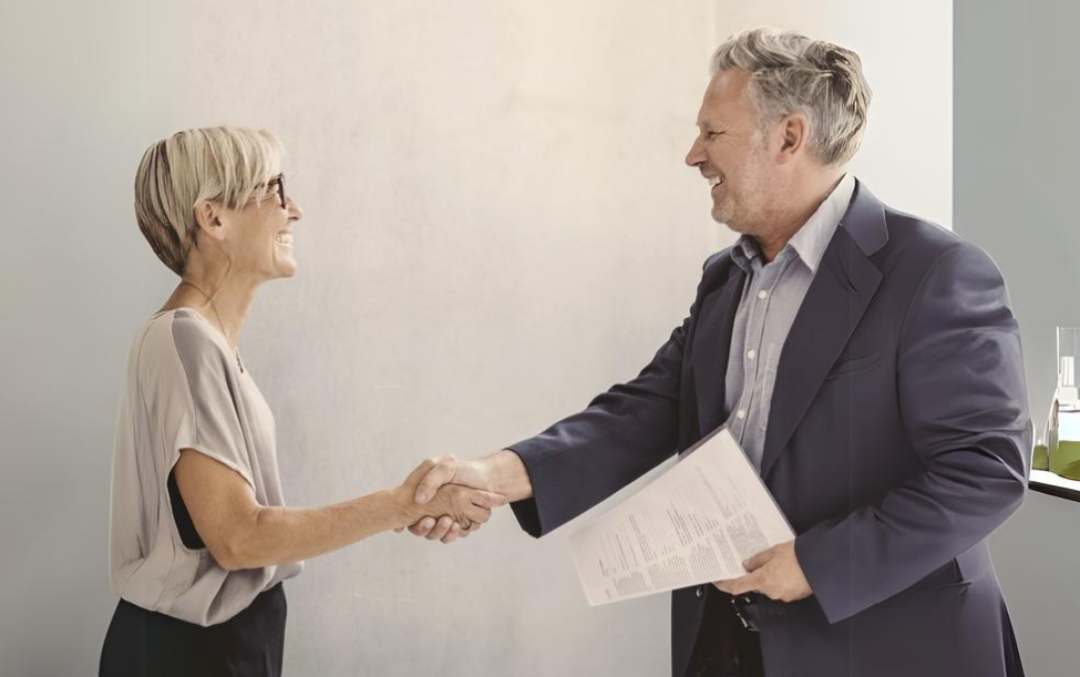 Zwei Personen schütteln sich die Hände. Die Person links trägt ein helles Oberteil und eine dunkle Hose, während die Person rechts einen dunklen Anzug und ein helles Hemd trägt und Dokumente in der linken Hand hält.
