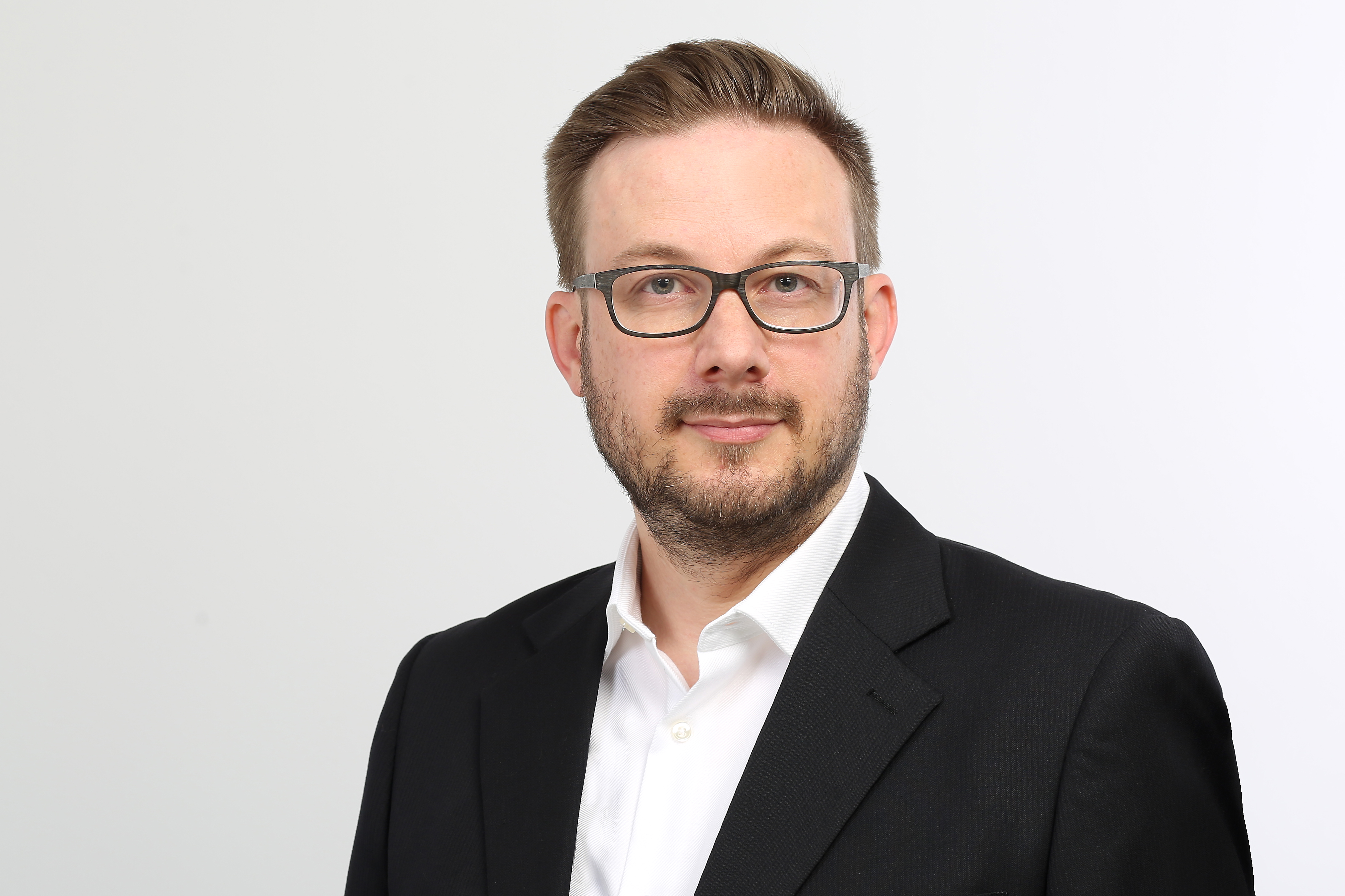 Leo Molatore, ein Mann in einem schwarzen Anzug und weißem Hemd, steht vor einem weißen Hintergrund.