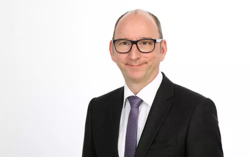 Mario Hartman, vom Management Team, ist in einem schwarzen Anzug mit weißem Hemd und lila Krawatte und steht vor einem weißen Hintergrund.