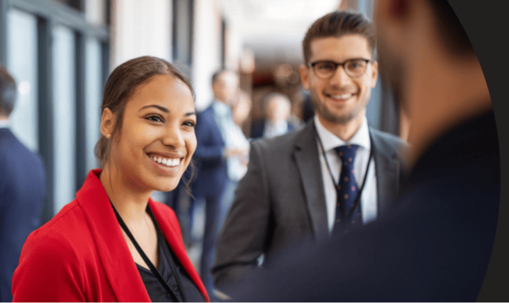 Gruppe von Personen auf einer geschäftlichen Veranstaltung, im Vordergrund drei Personen – eine im roten Blazer, eine im Anzug mit Krawatte, eine von hinten sichtbar; Gesichter unscharf.