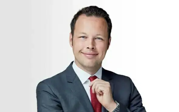 Simon Foelting trägt einen dunklen Anzug, ein weißes Hemd und eine rote Krawatte.