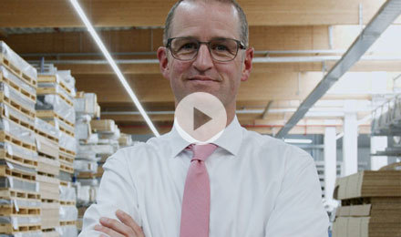 Eine Person steht mit verschränkten Armen in einem Lager oder Archiv. Sie trägt ein weißes Hemd und eine rosa Krawatte. Im Hintergrund sind Regale mit verschiedenen Gegenständen, möglicherweise Kartons oder Akten, zu sehen.