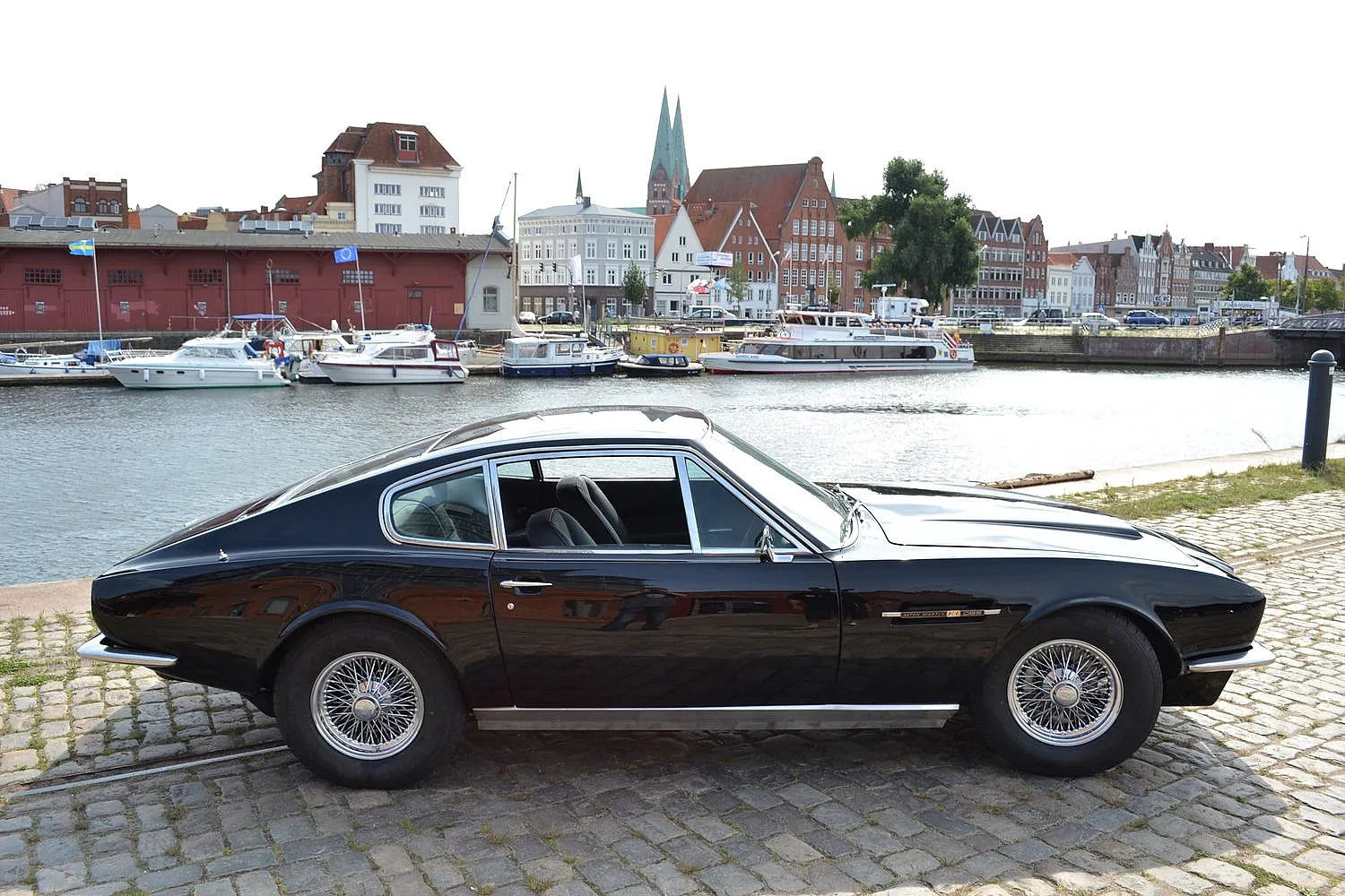 Schwarzer Aston Martin geparkt an einem Flussufer mit historischen Gebäuden und Booten im Hintergrund