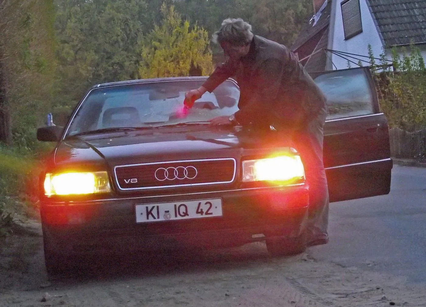 Jens Tanz reinigt die Windschutzscheibe seines schwarzen Audi v8 bei der Dämmerung. Die Scheinwerfer sind auch und das Auto parkt am Straßenrand.