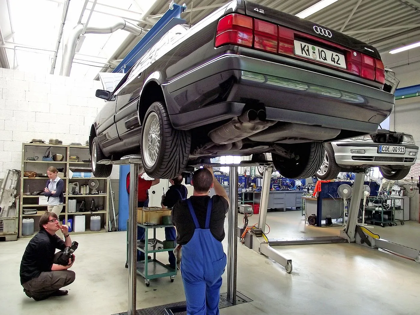 Blick unter den Audi v8 in der Werkstatt auf einer Hebebühne. Zwei Männer begutachten den Wagen von unten.
