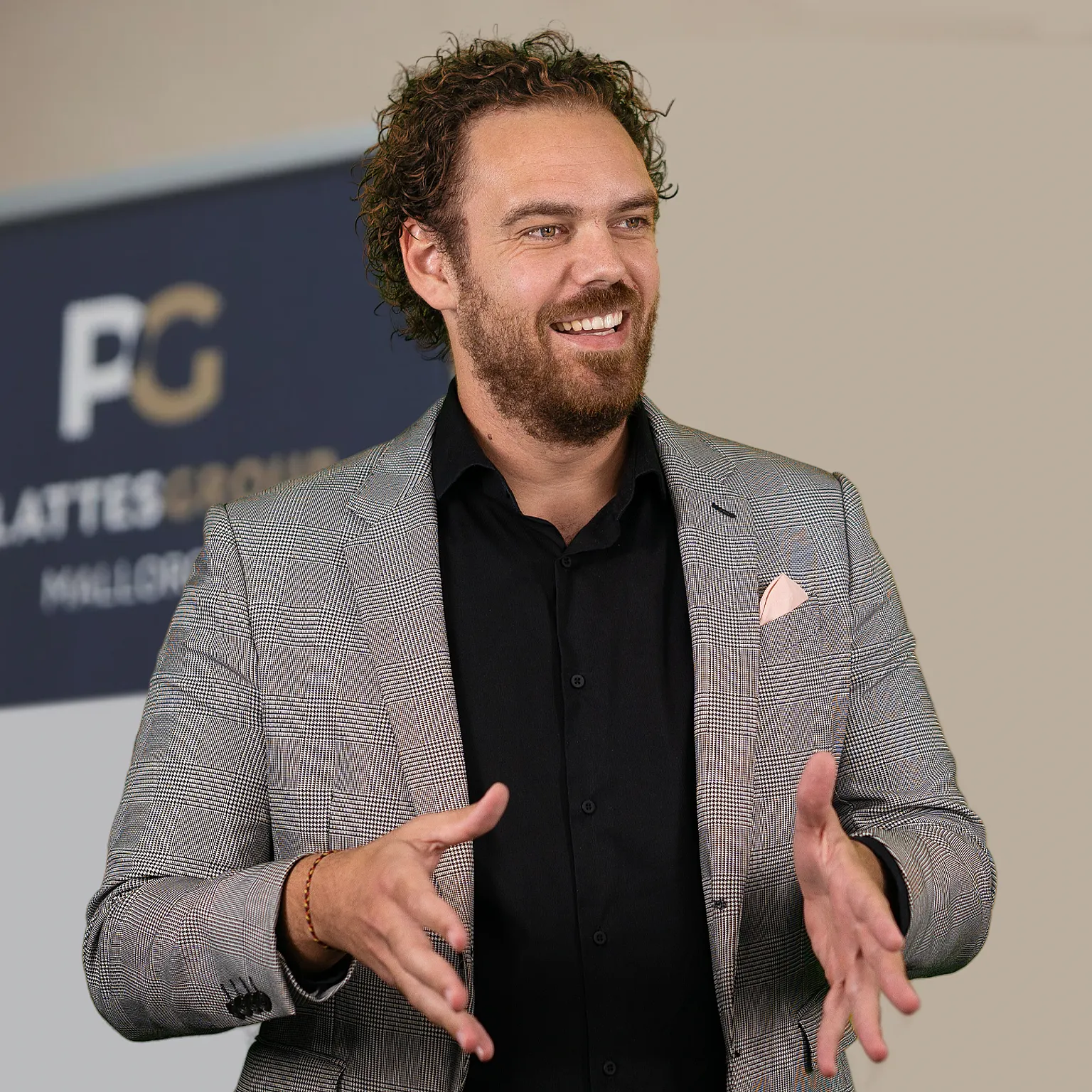 Christian Plattes mit lockigem Haar und Bart, in einem grauen Anzug und schwarzem Hemd, lächelnd und gestikulierend. Im Hintergrund ist ein unscharfes Schild mit dem „PG“-Logo zu sehen.