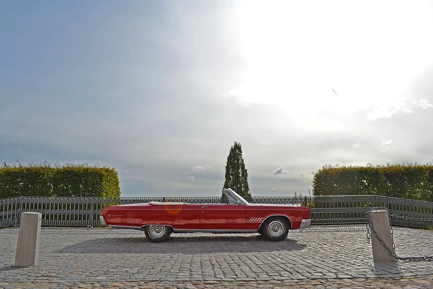 Ein rotes Cabriolet steht auf einem gepflasterten Platz vor einem Zaun und hohen Hecken. Der Himmel ist bewölkt, aber hell