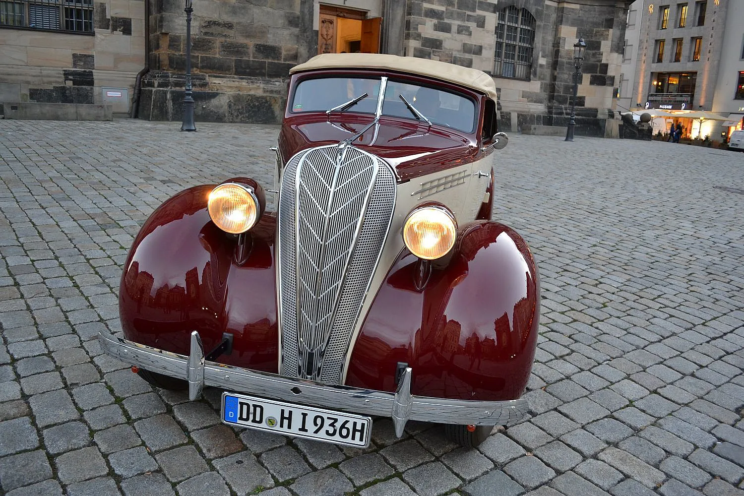 Ein Oldtimer ist auf einer Kopfsteinpflasterstraße vor einem alten Gebäude geparkt. Das Auto hat ein markantes, klassisches Design mit einem auffälligen Kühlergrill und runden Scheinwerfern.