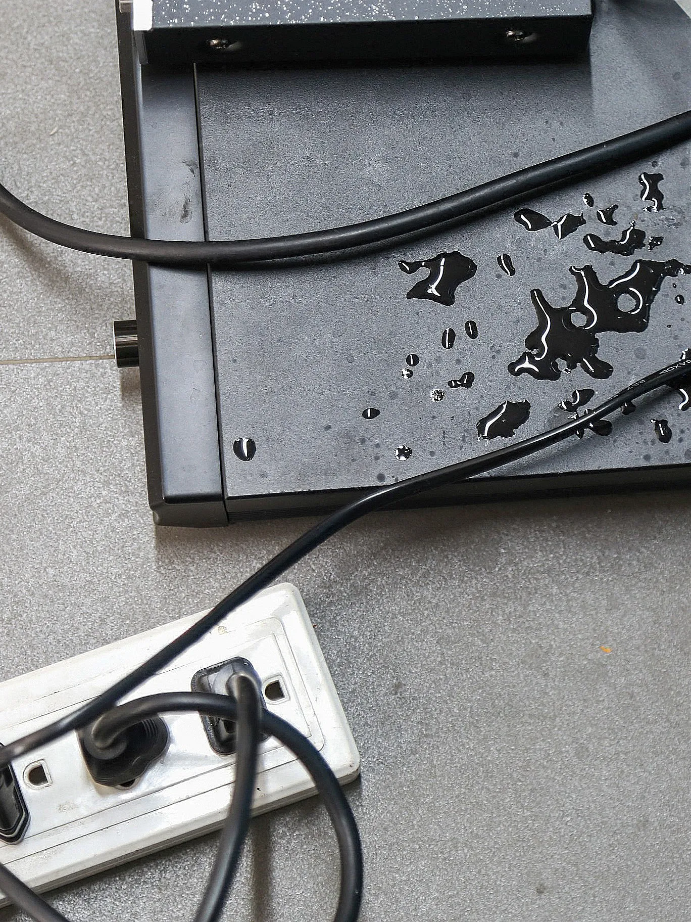 Nasse Stromkabel und eine Steckdosenleiste auf grauem Boden, mit Wassertropfen auf einem schwarzen Gerät, was eine gefährliche Situation darstellt.