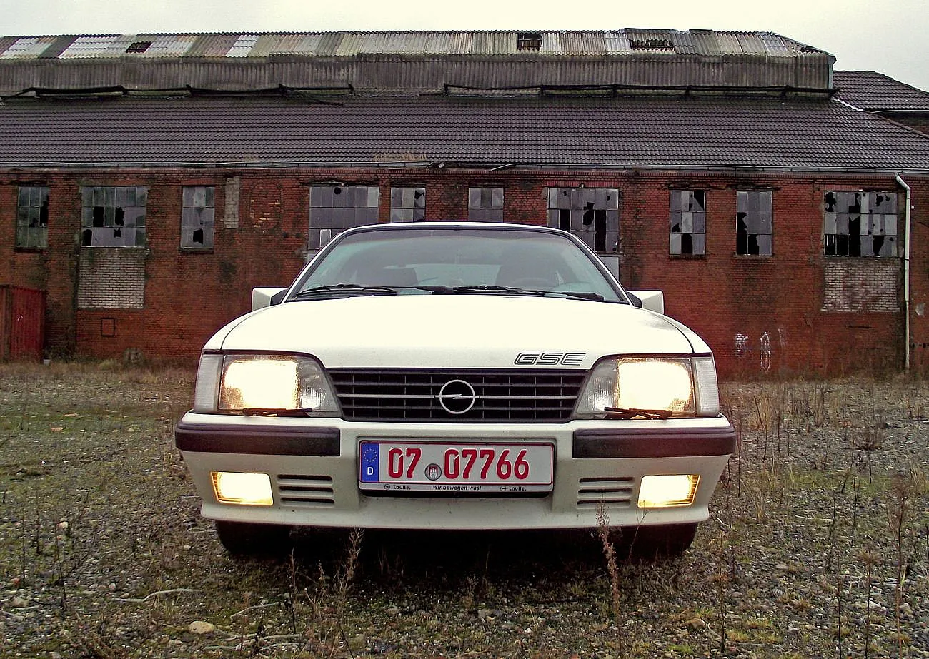 Weißer Opel mit eingeschalteten Scheinwerfern, geparkt vor einem verlassenen Backsteingebäude mit zerbrochenen Fenstern.