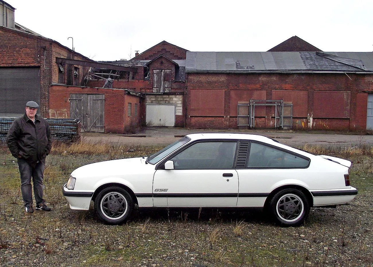 Weißer parkender Opel Monza GSE mit Seitenansicht vor einem Haus daneben steht ein Mann in Jacke und Hut.