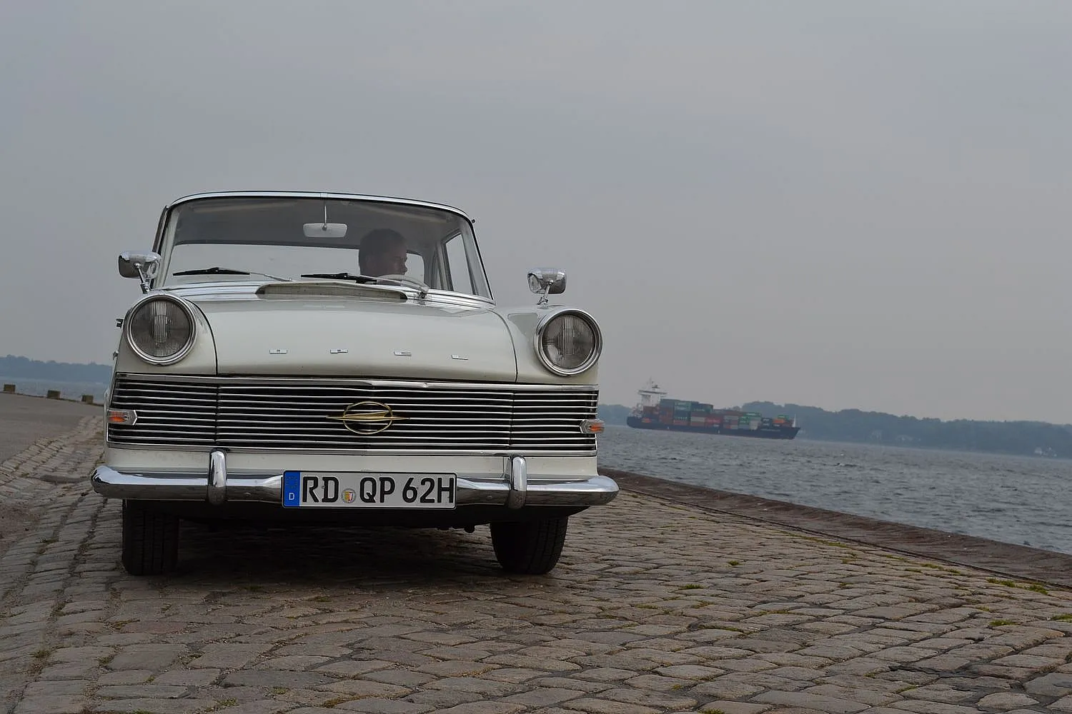  Weißes klassisches Auto mit dem Kennzeichen "RD QP 62H" auf einer Kopfsteinpflasterstraße nahe einem Gewässer, im Hintergrund ein großes Frachtschiff.