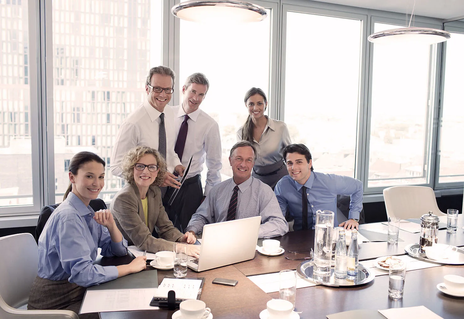 Sieben lächelnde Geschäftsleute sitzen um einen Laptop an einem Meetingtisch in Business-Kleidung.