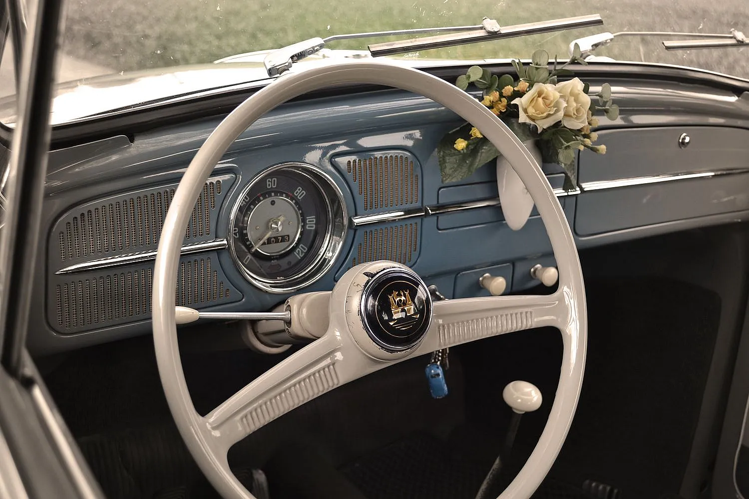 Innenraum eines Oldtimers mit weißem Lenkrad, blauem Armaturenbrett und einem kleinen Blumenstrauß auf dem Armaturenbrett. Ein blauer Schlüssel hängt am Lenkrad.