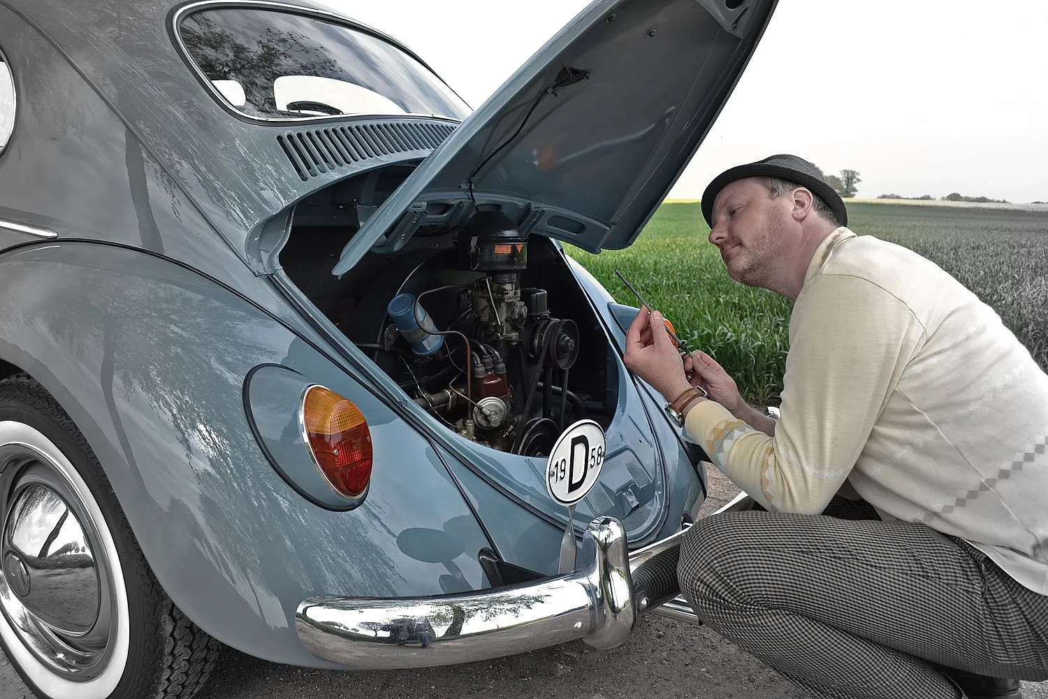 Eine Person arbeitet am Motor eines klassischen VW Käfers mit geöffneter Heckklappe. Die Person kniet neben dem Auto und benutzt Werkzeuge.
