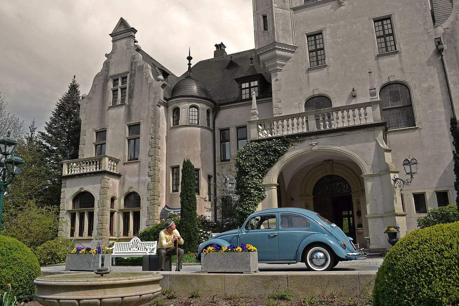 Ein historisches Gebäude mit Steinmauern, Rundbogenfenstern und einem Turm. Davor steht ein blauer Oldtimer, eine weiße Bank und eine Person mit Hut in heller Kleidung.