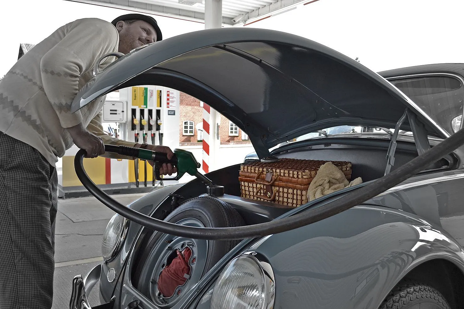 Eine Person tankt einen Oldtimer an einer Tankstelle. Die Motorhaube ist geöffnet und zeigt den Motor sowie einen Weidenkorb. Im Hintergrund sind mehrere Zapfsäulen zu sehen.