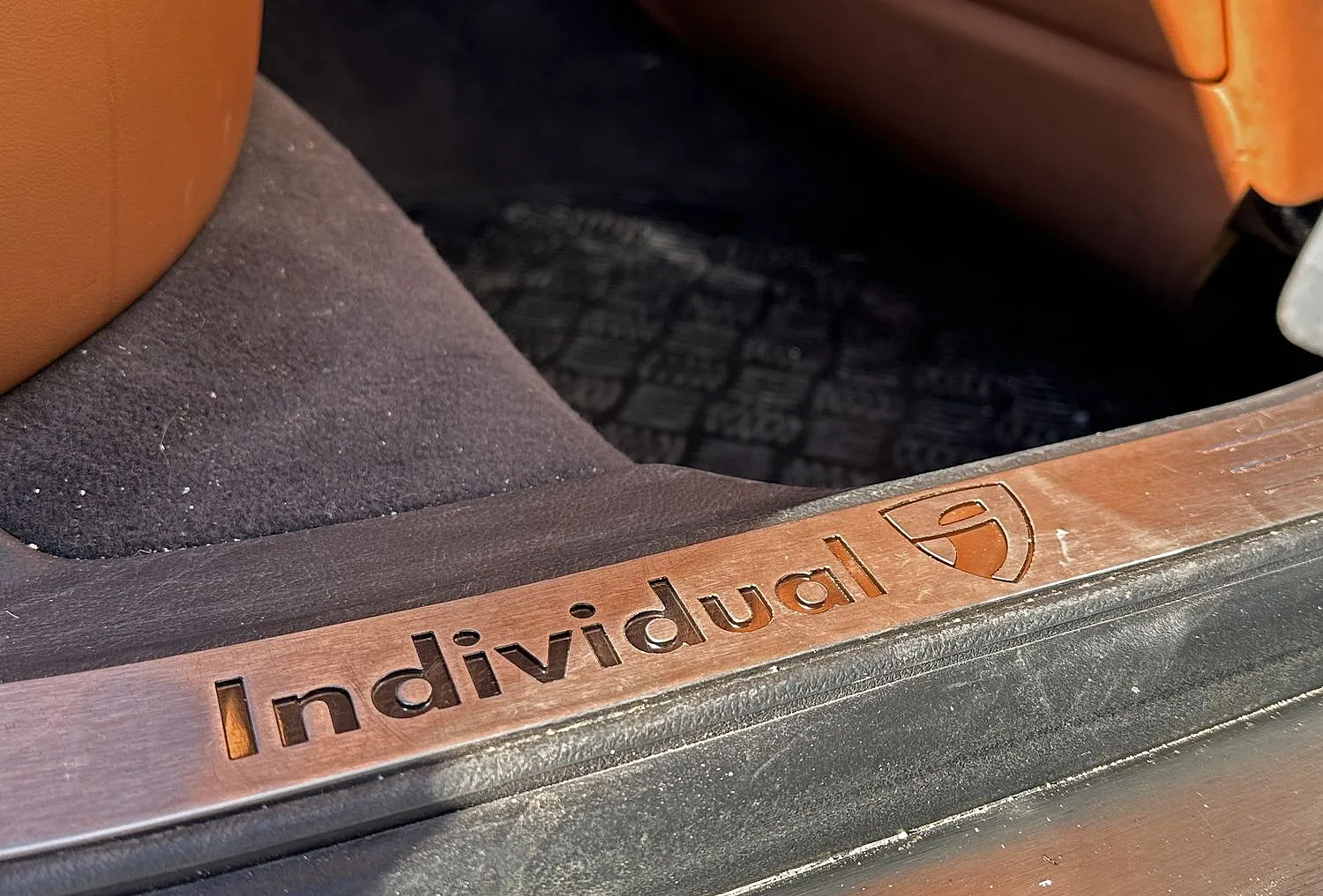 Nahaufnahme einer Autotürschwelle mit der Gravur „Individual“ auf einem Metallschild. Braune Ledersitze und ein dunkler Teppich sorgen für ein luxuriöses Gefühl.