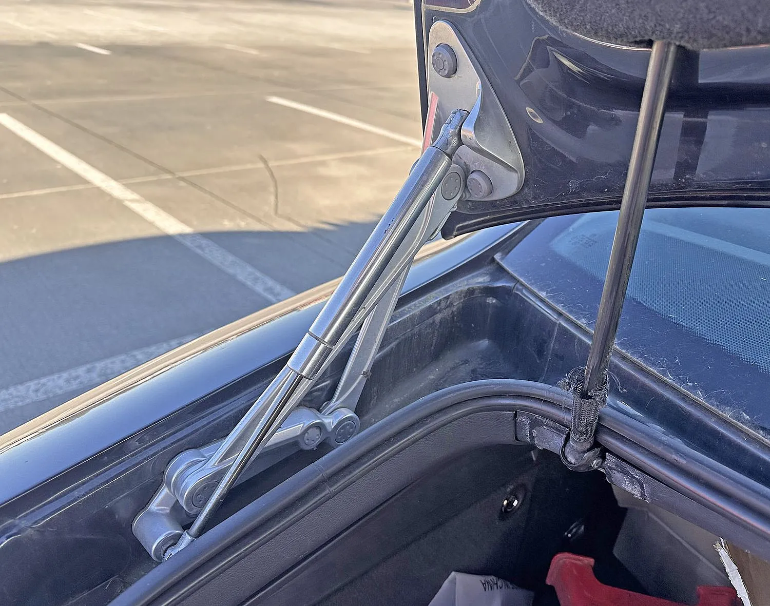 Nahaufnahme eines Kofferraumscharniers und einer Gasdruckfeder, teilweise geöffnet. Das Sonnenlicht hebt die Metallteile hervor, im Hintergrund ist ein Parkplatz zu sehen.