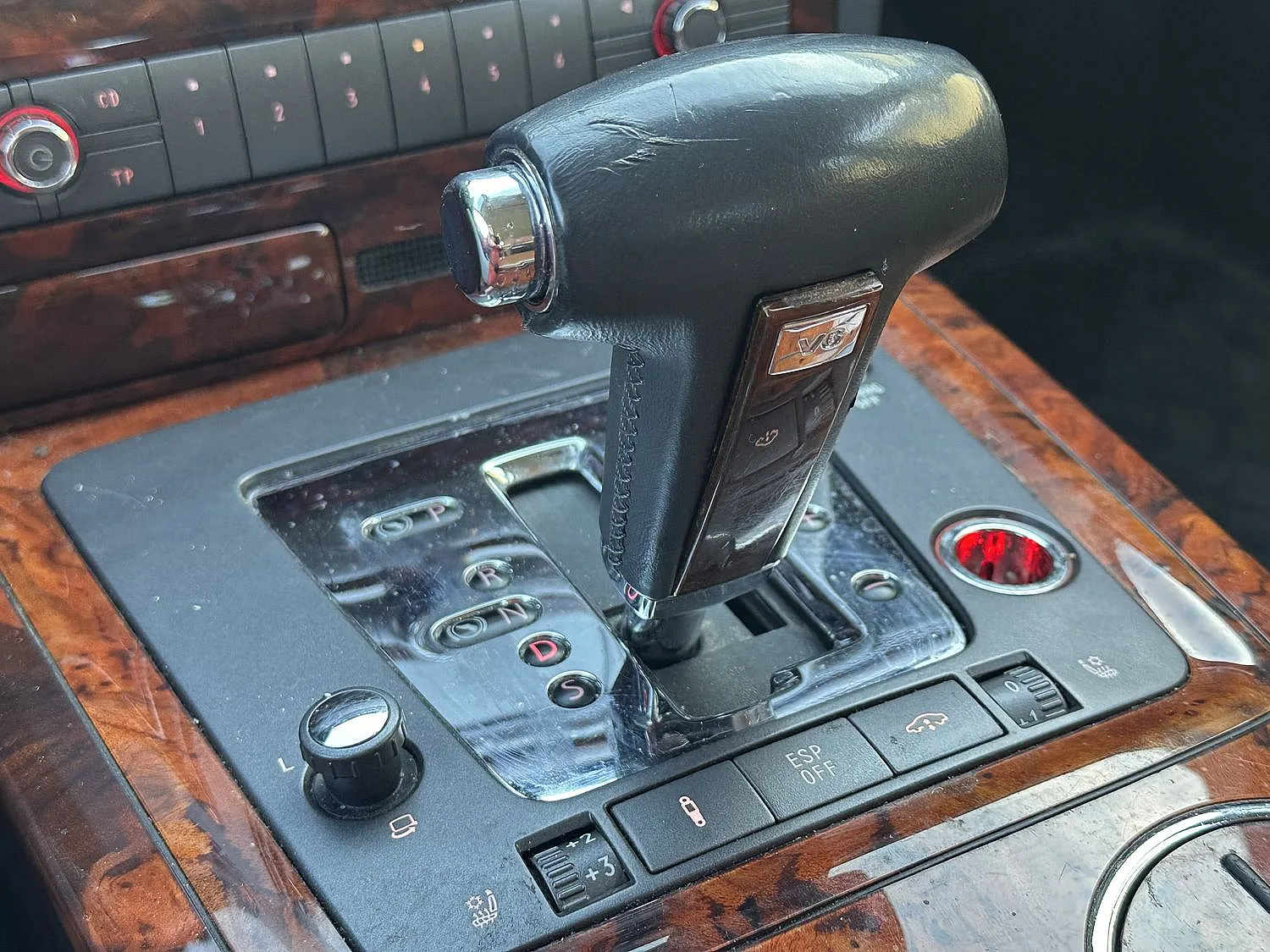 Nahaufnahme des Schalthebels eines VW Phaeton aus schwarzem Leder auf einer polierten Konsole in Holzoptik, mit einem V6-Emblem und detaillierten Bedientasten.