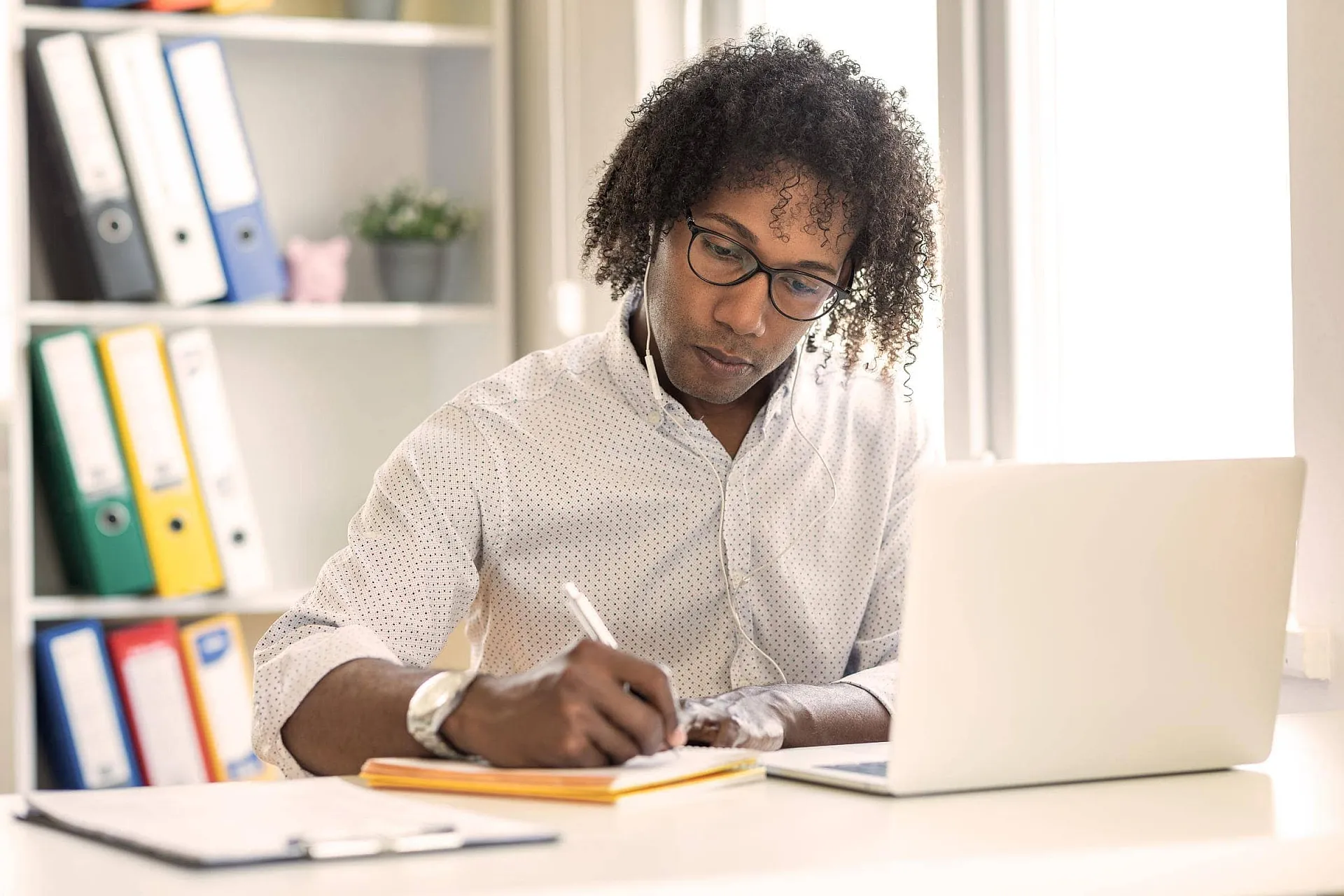 Ein Blogger mit Brille schreibt auf einem Blatt Papier, während er Kopfhörer trägt und an seinem Laptop arbeitet.