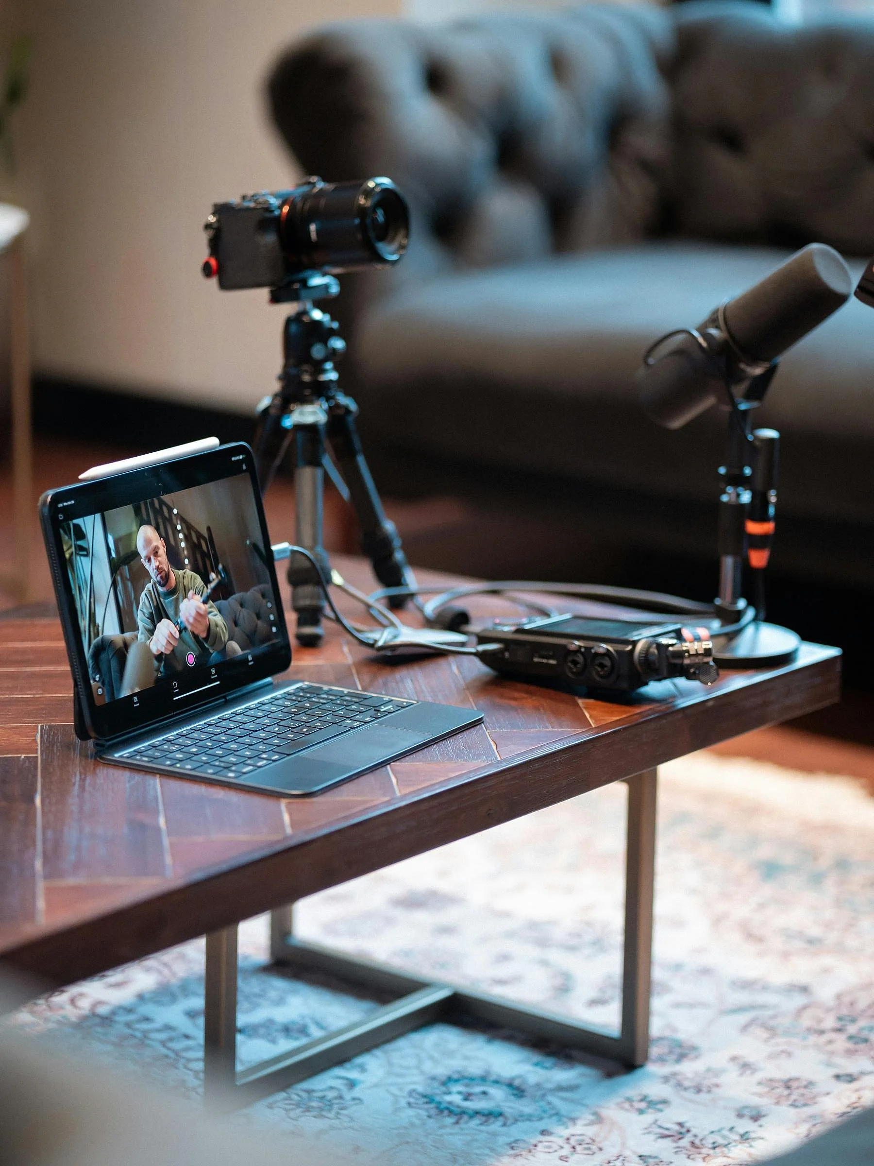 Ein Podcast-Setup zu Hause auf einem Holztisch, bestehend aus einem Laptop, auf dem ein Interview zu sehen ist, einer Kamera auf einem Stativ, Mikrofonen und Audiogeräten. Gemütliche Atmosphäre.