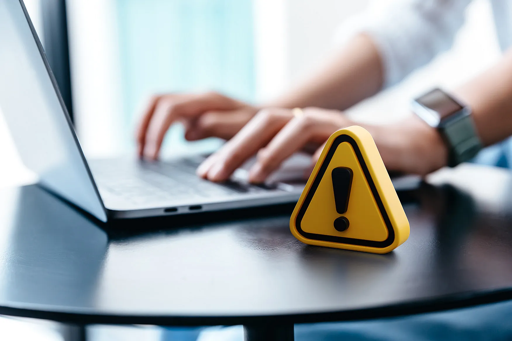 Person, die an einem Tisch auf einem Laptop tippt, mit einem gelben Warndreieck mit Ausrufezeichen im Vordergrund, das zur Vorsicht oder Wachsamkeit mahnt.