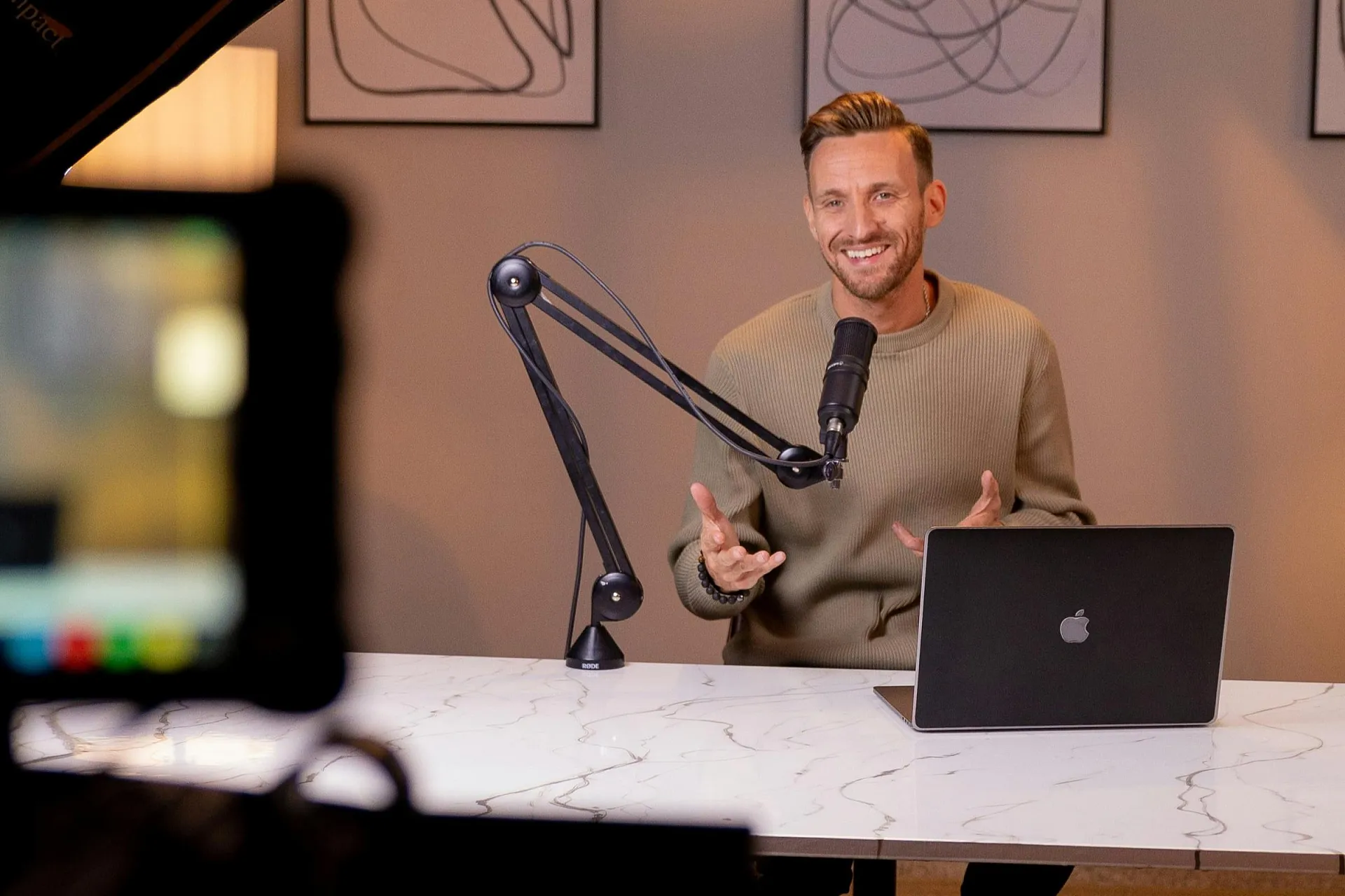 Ein Mann, der während einer Podcast-Aufnahme lächelt und in ein Mikrofon spricht. Er sitzt an einem Marmortisch mit einem Laptop, im Hintergrund sind gerahmte Bilder zu sehen.