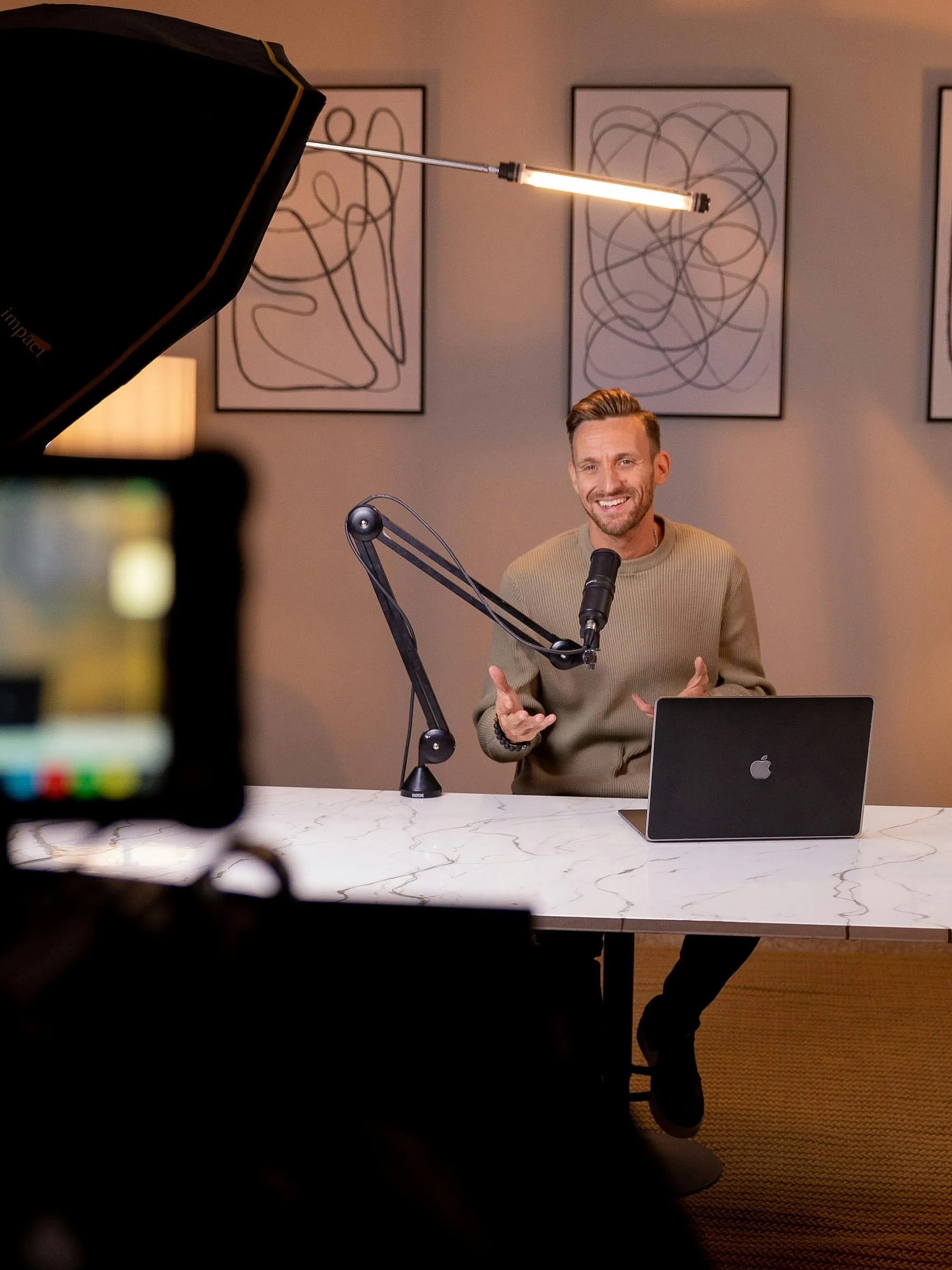 Ein Mann, der während einer Podcast-Aufnahme lächelt und in ein Mikrofon spricht. Er sitzt an einem Marmortisch mit einem Laptop, im Hintergrund sind gerahmte Bilder zu sehen.