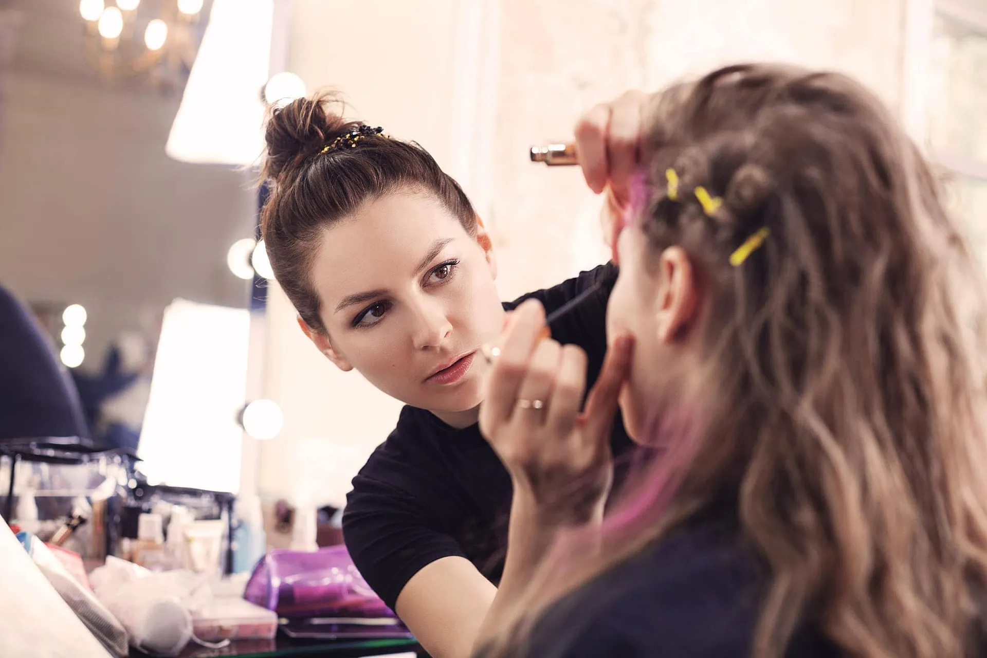 Beauty-Versicherung: Make-Up-Artist mit hochgebundenen Haaren trägt einer Kundin Wimperntusche auf