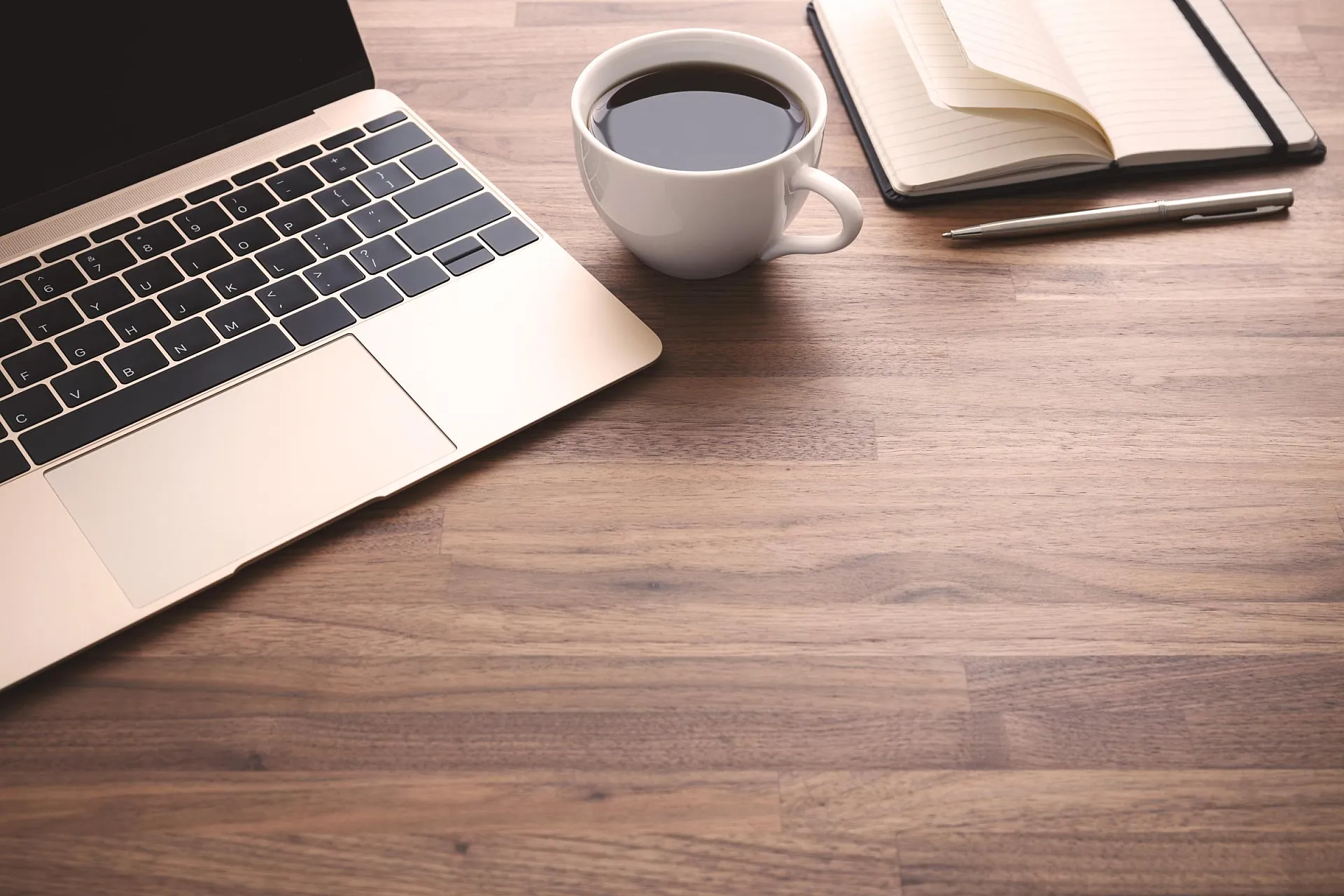 Auf einem hellbraunen Holztisch stehen ein Laptop, eine Tasse Kaffee, ein Stift und ein Notizbuch.