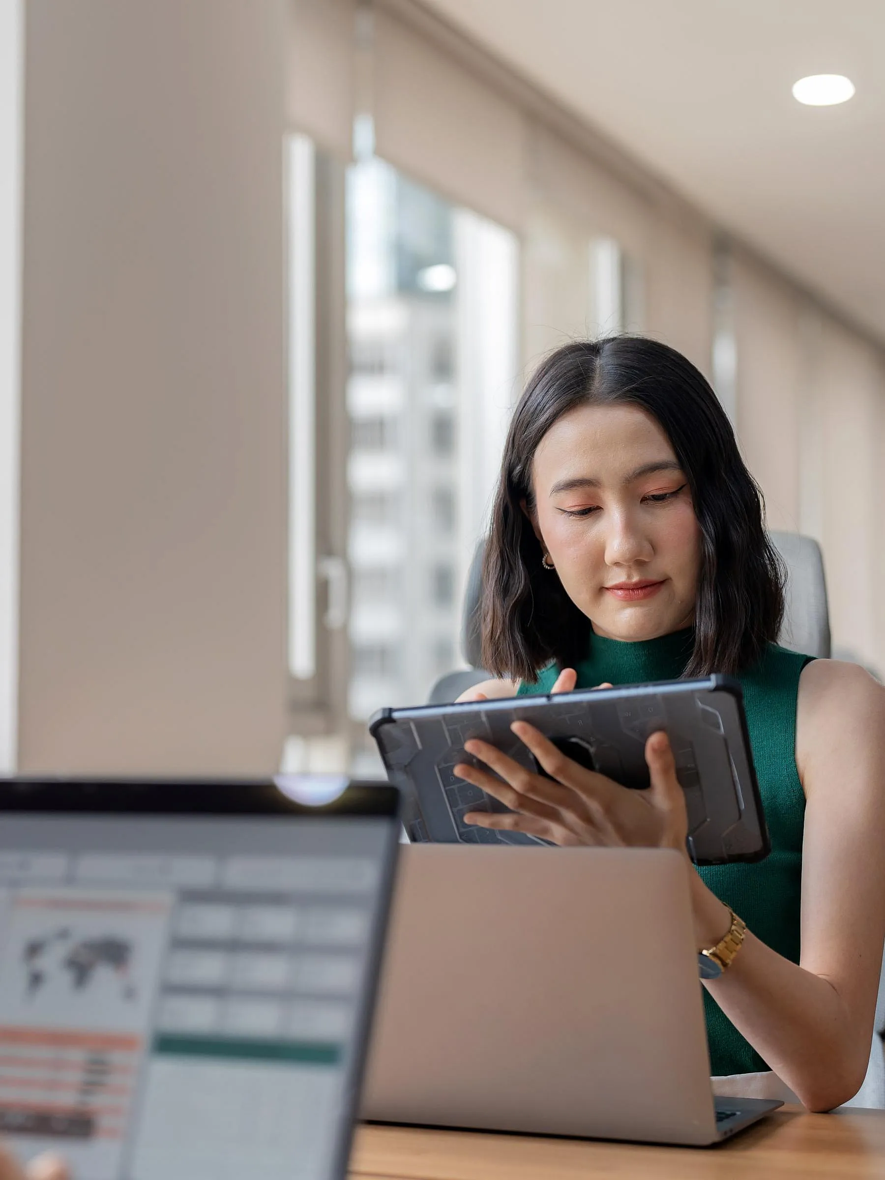 In einem hellen Büro arbeitet eine Frau in grüner Kleidung konzentriert an einem Tablet, während im Vordergrund ein Mann an einem Laptop mit Grafiken zu sehen ist, dessen Bild verschwommen ist.