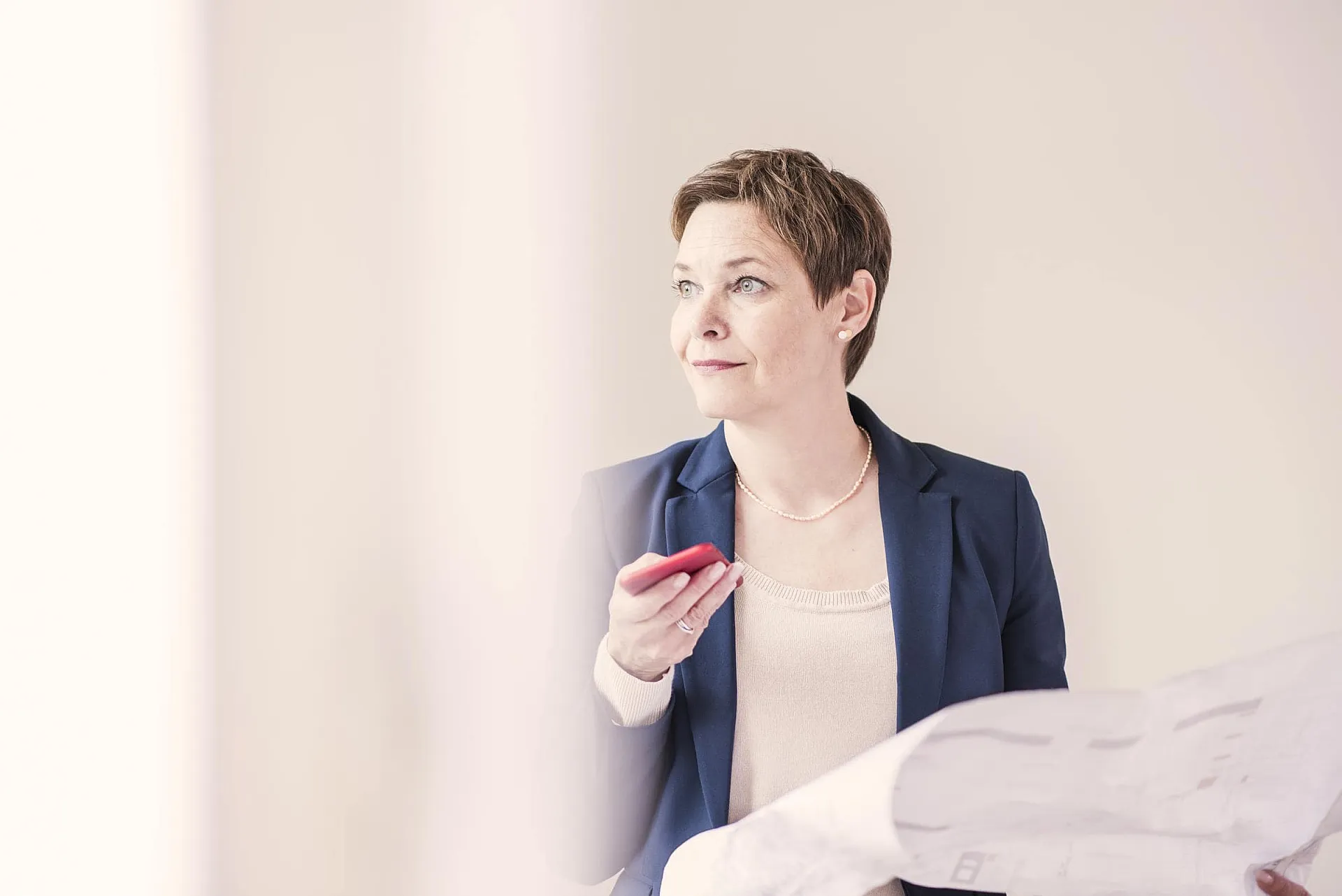Eine Frau im Businessanzug hält in einer Hand ein Mobiltelefon mit einer roten Handyhülle und in der anderen ein großen Zettel und schaut dabei seitlich in die Ferne.