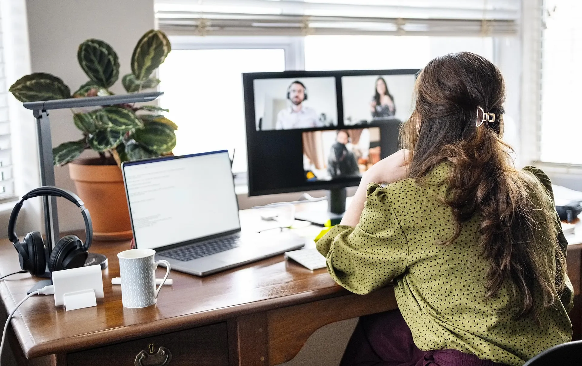 Eine Frau in einer grünen Bluse arbeitet von zu Hause aus und nimmt an einer Videokonferenz auf ihrem Computer teil. Sie sitzt an einem Schreibtisch, auf dem ein Laptop, Kopfhörer und eine Topfpflanze stehen.