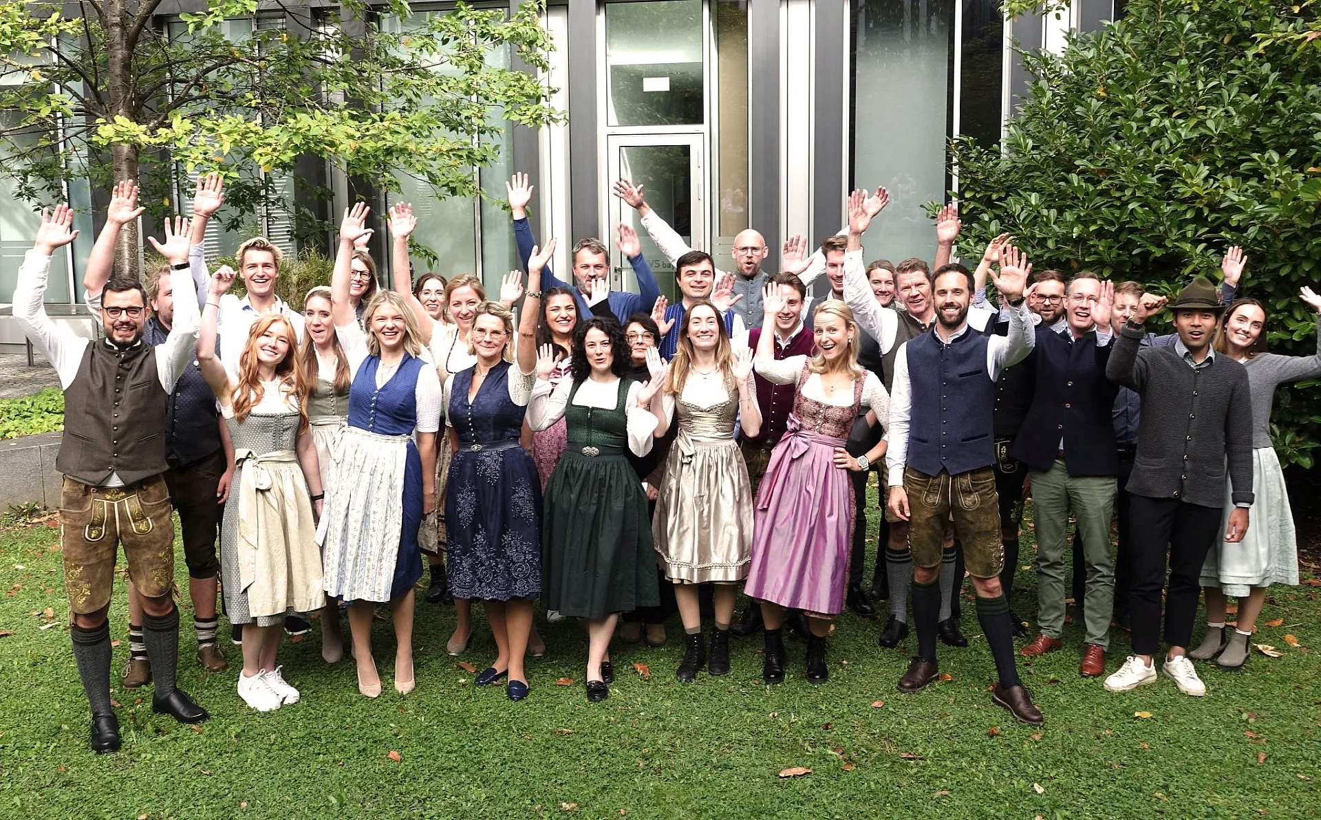 Die Mitarbeiter von Hiscox München stehen in einer großen Gruppe in bayrischer Tracht im Innenhof des Münchner Büros auf der grünen Wiese. Alle haben die Hände und lachen in die Kamera.