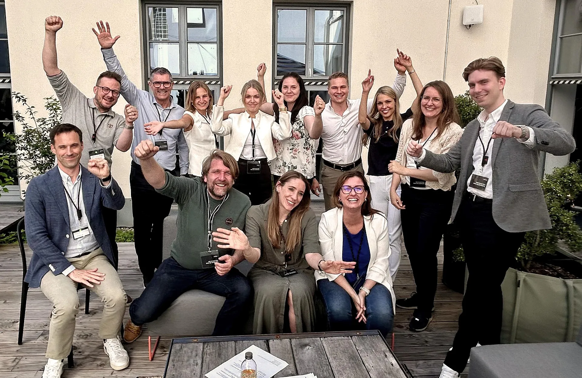Eine Gruppe von fünfzehn Personen posiert auf einer Terrasse für ein Foto. Einige sitzen auf Sofas, während andere stehen. Alle haben ihre Gesichter verdeckt und tragen Namensschilder um den Hals. Die Stimmung scheint fröhlich und ausgelassen zu sein, da viele Personen ihre Arme in die Luft strecken oder Daumen hoch zeigen