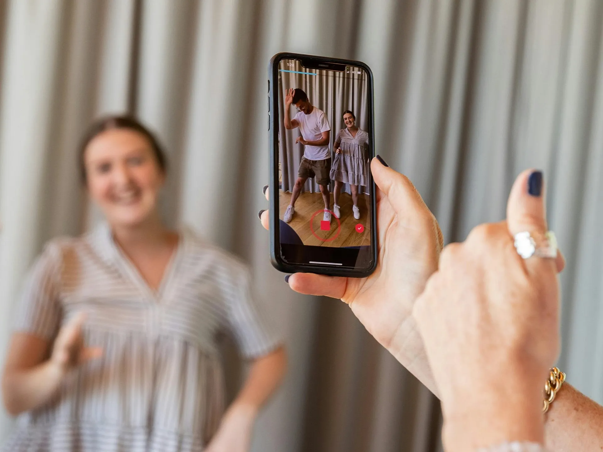 Eine Person hält ein Handy in der Hand und filmt zwei Menschen, die in einem Raum tanzen. Die Frau im Fokus trägt ein gestreiftes Kleid und wirkt vor dem Hintergrund eines verschwommenen Vorhangs fröhlich.