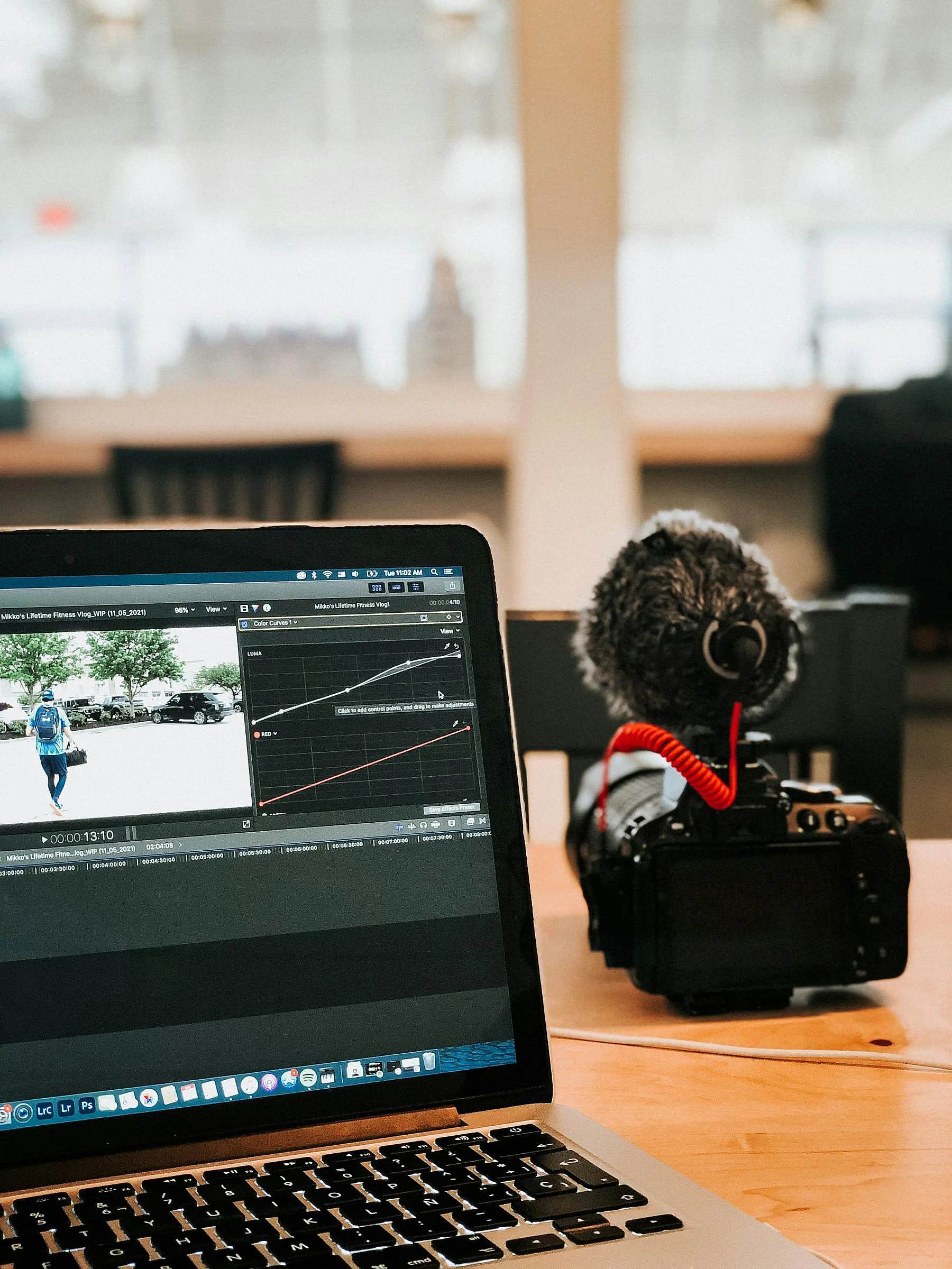 Auf einem Holztisch steht ein Laptop, auf dem eine Videobearbeitungssoftware läuft, daneben eine Kamera mit Mikrofon. Die Szene vermittelt den Eindruck eines kreativen Arbeitsbereichs.