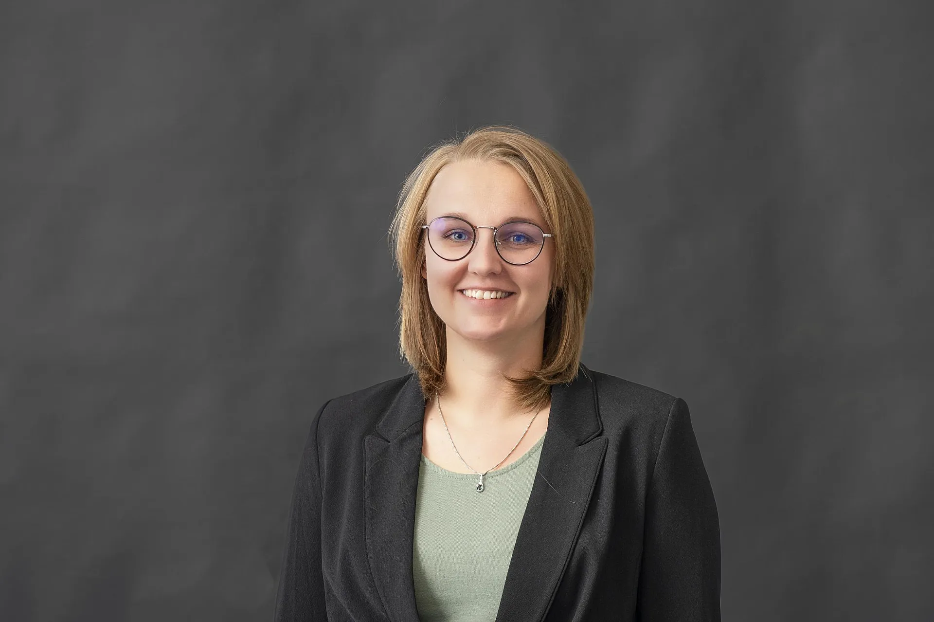 Milena Leininger trägt einen schwarzen Blazer über einem grünen Oberteil und steht vor einem dunklen Hintergrund.
