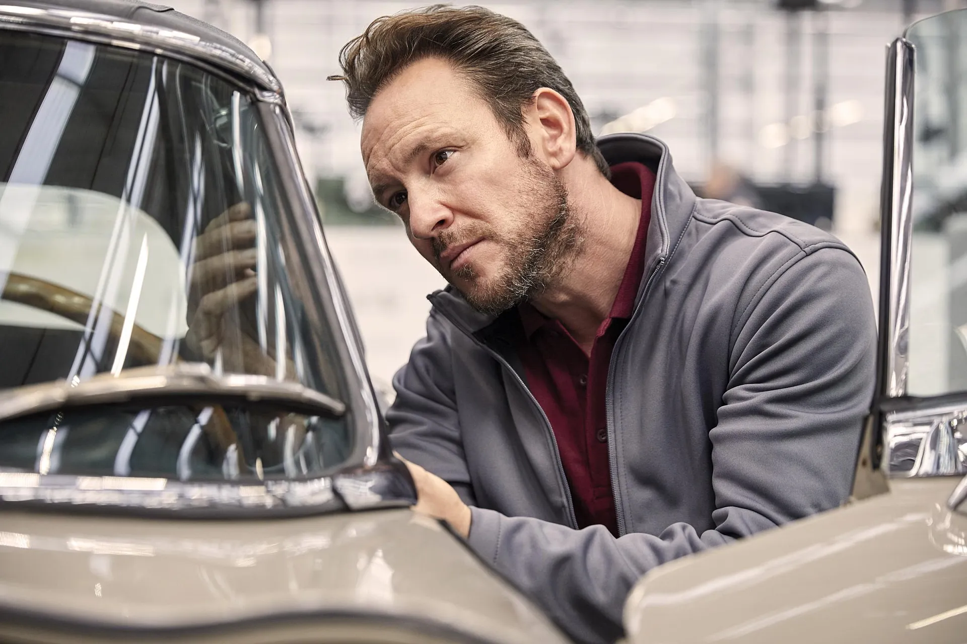 Eine Person mit verschwommenem Gesicht lehnt sich an das Fenster eines klassischen Autos. Die Person trägt eine graue Jacke über einem roten Hemd. Die Szene scheint sich in einer Ausstellung oder einem Showroom abzuspielen.