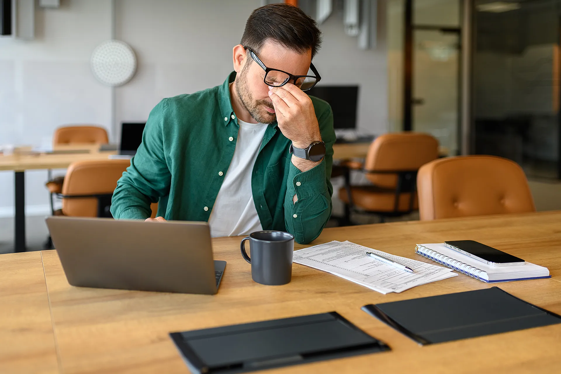 Ein Mann in einem grünen Hemd sitzt an einem Schreibtisch, sieht gestresst aus und drückt sich die Nasenwurzel. Er ist umgeben von Papieren, einem Laptop, einem Tablet und einer Kaffeetasse.