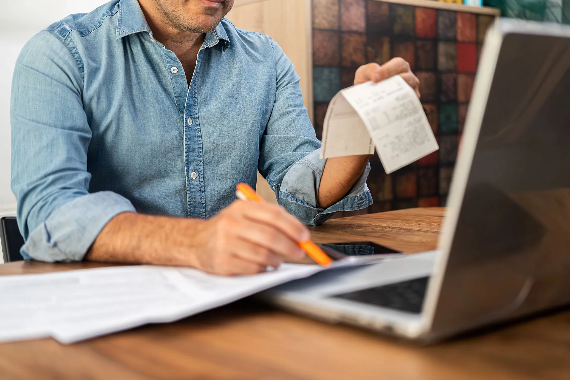 Ein Mann sitzt an einem Holztisch, trägt ein Jeanshemd und arbeitet am Laptop. In der einen Hand hält er einen Beleg oder eine Quittung, mit der anderen schreibt er etwas auf ein Blatt Papier. Auf dem Tisch liegen mehrere Dokumente.
