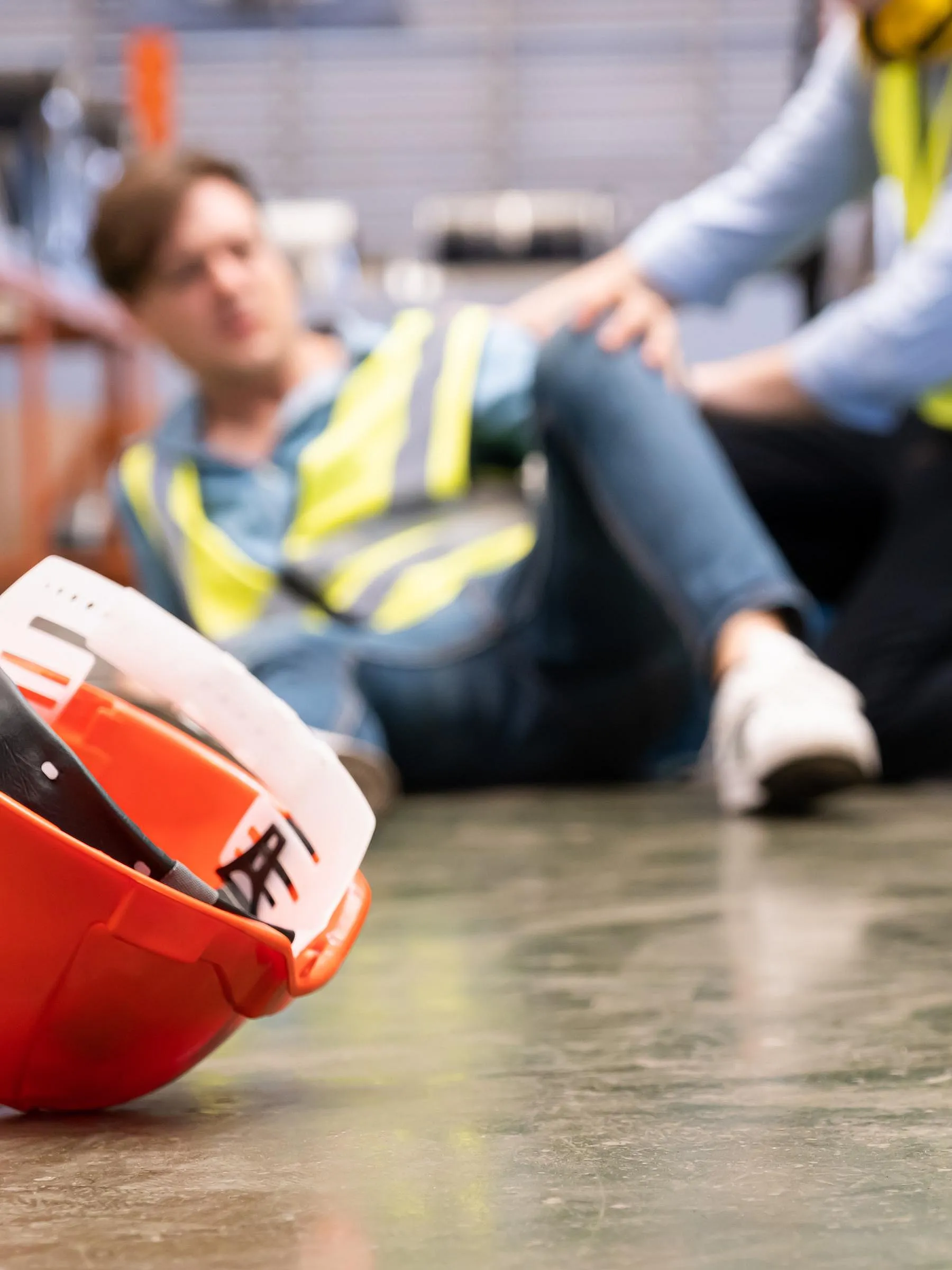 Ein roter Schutzhelm steht im Mittelpunkt auf dem Boden eines Lagerhauses. Im unscharfen Hintergrund kümmern sich zwei Arbeiter in Warnwesten um einen gestürzten Kollegen und zeigen ihre Besorgnis.