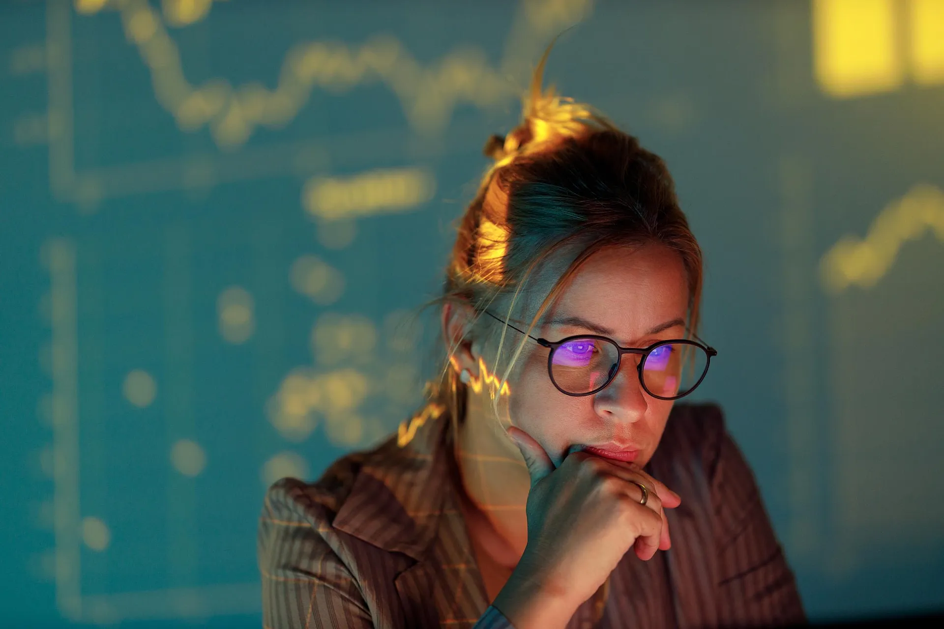 Eine Frau mit Brille blickt konzentriert auf einen Bildschirm, der von Finanzdiagrammen beleuchtet wird, die im Hintergrund projiziert werden. Die Szene strahlt eine nachdenkliche, analytische Atmosphäre aus.