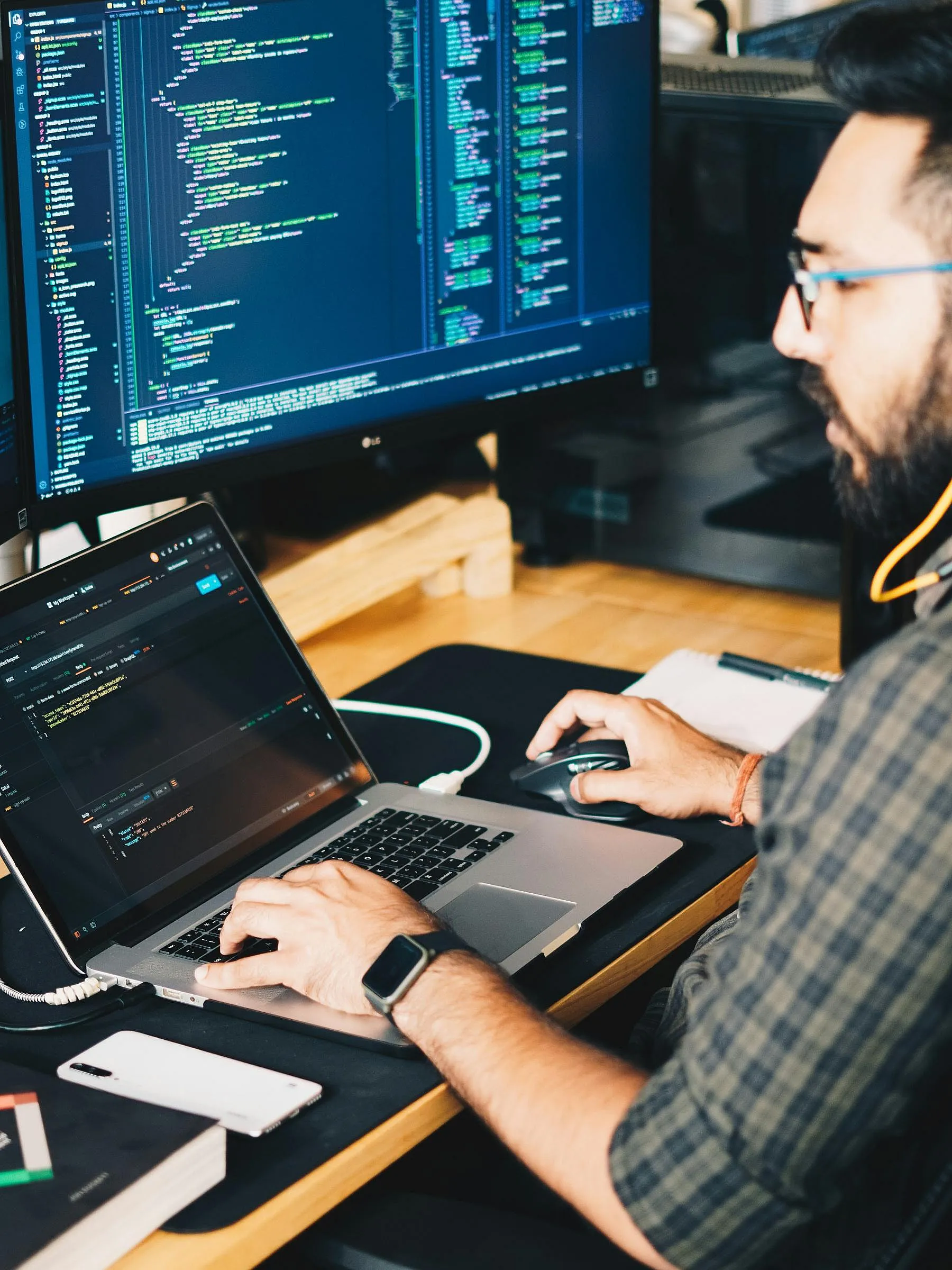 Ein Mann sitzt an einem Schreibtisch mit einem Laptop und einem großen Monitor, auf dem Code angezeigt wird. Er wirkt konzentriert, bedient die Maus und befindet sich in einer von Technik und Produktivität geprägten Umgebung.